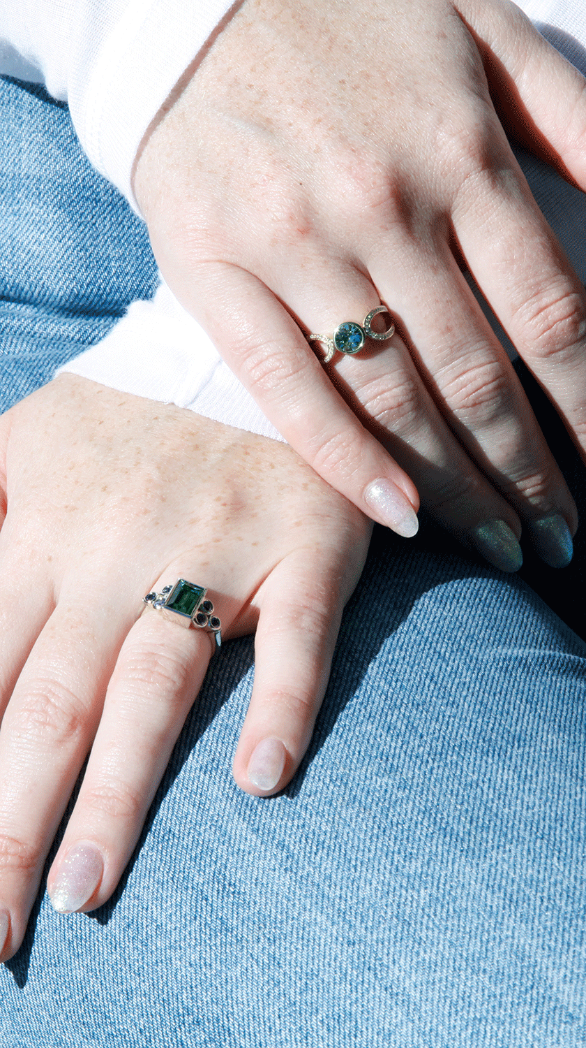 A close-up of hands adorned with two unique gemstone rings, perfect for casual or formal wear. One hand showcases a green rectangular gemstone set in a silver band with intricate detailing, while the other features a celestial-inspired ring with a round b