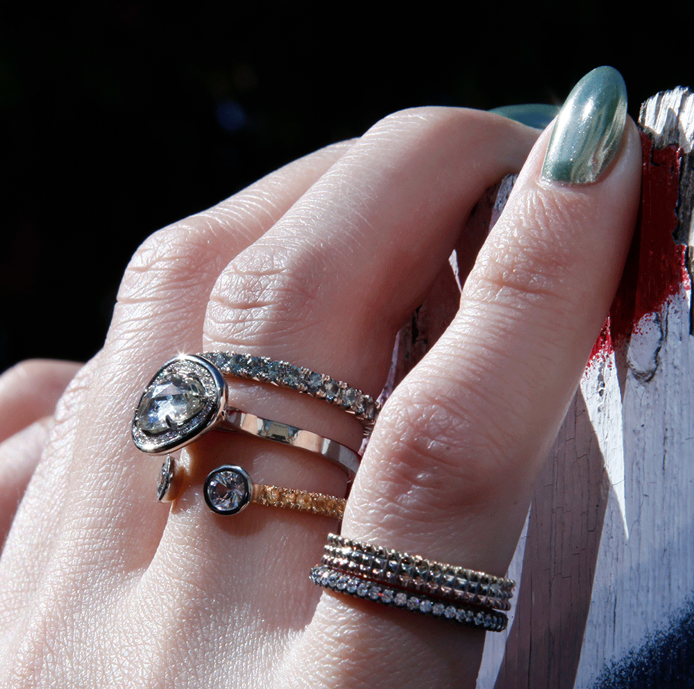 The rings appear to be of varying styles and sizes. The nails are painted a light color. The background is dark and out of focus.