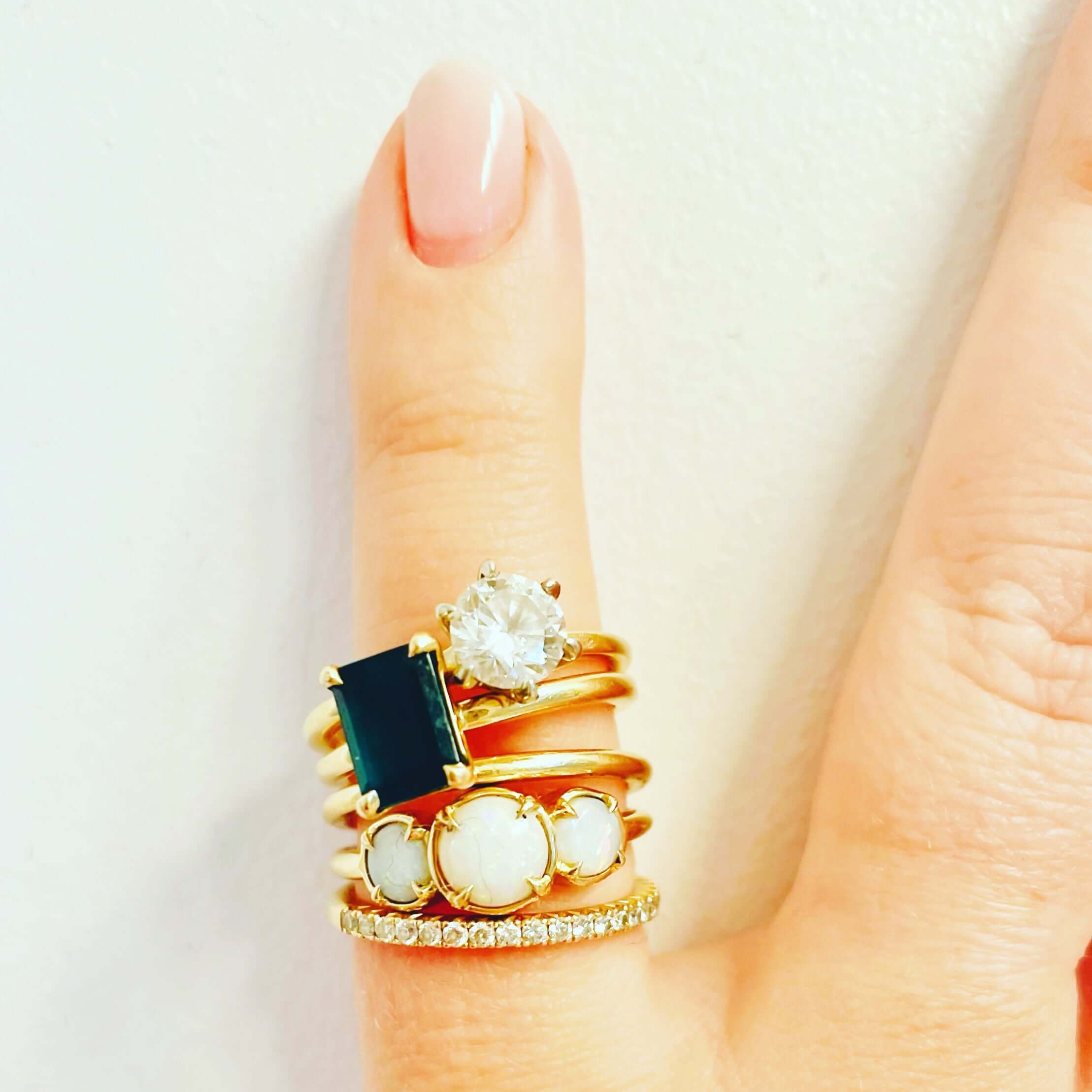 A close-up of a hand adorned with stacked gold rings featuring opals, diamonds, and a deep green gemstone. A delicate and meaningful jewellery collection symbolizing love, new beginnings, and cherished milestones.