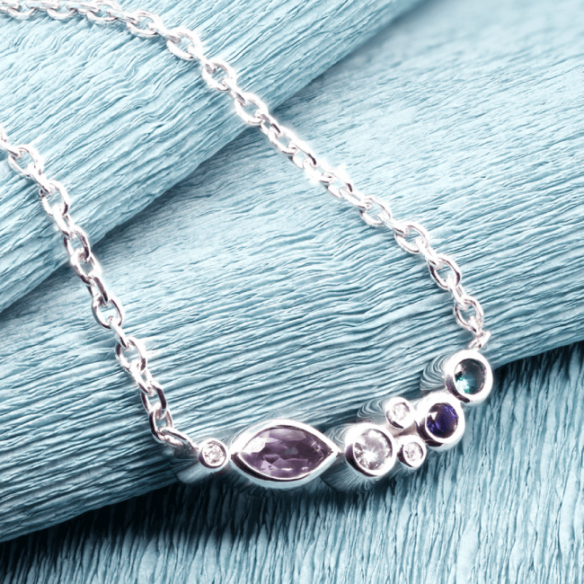 A delicate silver necklace or bracelet with small, possibly purple or reddish-purple, gemstones resting on a light teal or blue fabric background.