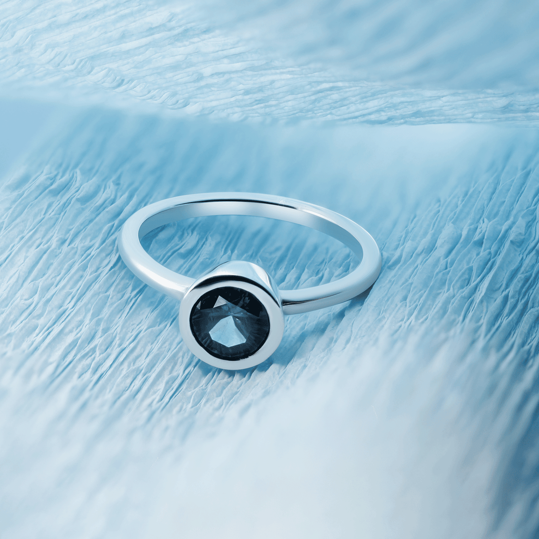 Single round blue sapphire solitaire ring set in gleaming 18K recycled white gold, photographed on a clean blue background. Front view