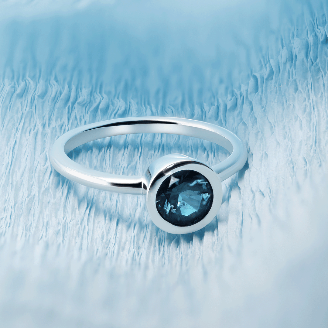 Single round blue sapphire solitaire ring set in gleaming 18K recycled white gold, photographed on a blue background. Front view