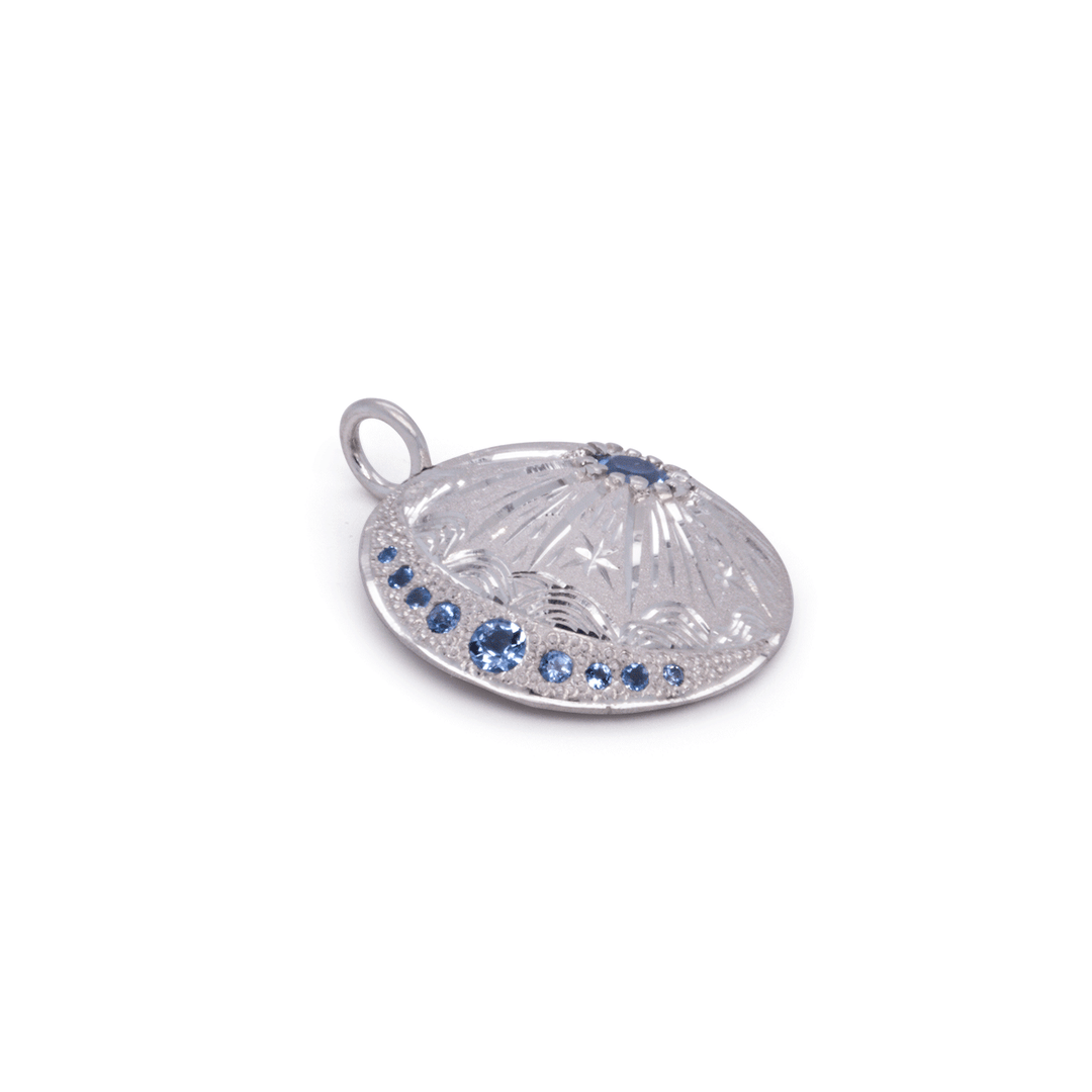 Handcrafted silver starburst charm pendant with celestial engraving and sparkling blue topaz accents by Janine Jewels.