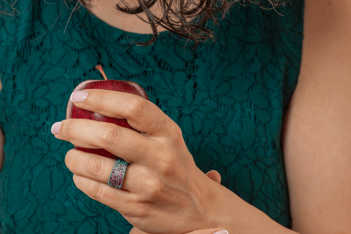 woman holding an apple, wearing a titanium ring fully paved in mostly pink lab sapphires. Titanium is a teal colour.