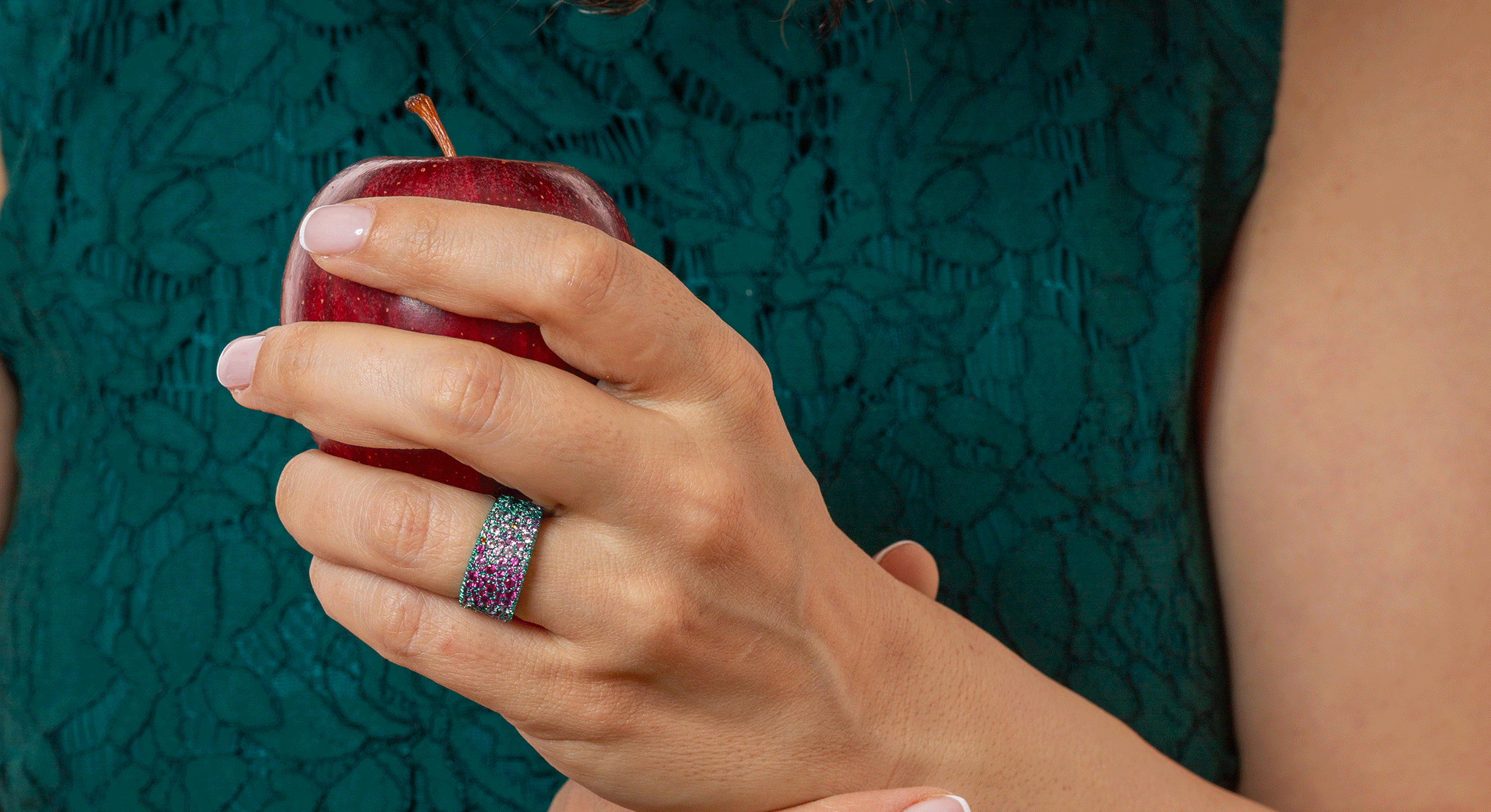 woman holding an apple, wearing a titanium ring fully paved in mostly pink lab sapphires. Titanium is a teal colour.