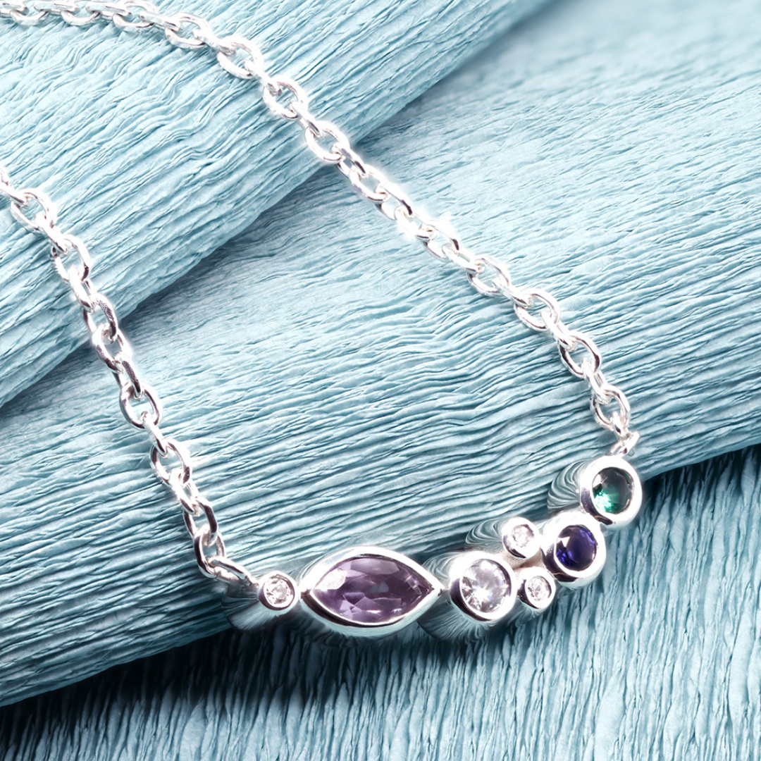 A close-up of a silver necklace featuring a marquise-cut purple gemstone and a combination of smaller round gemstones in various colours, elegantly displayed against a textured light blue background.