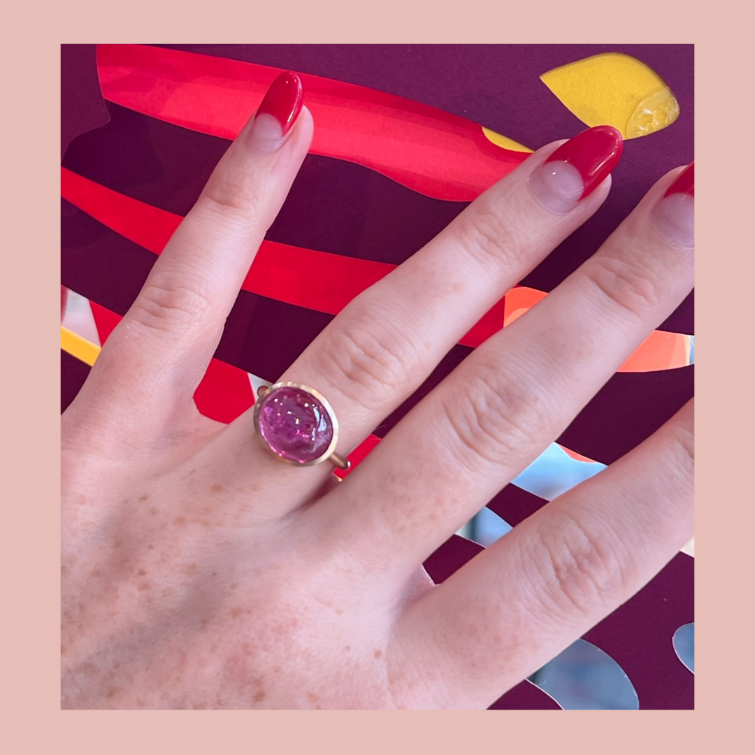 A romantic Valentine’s Day-themed image featuring  a close-up of a hand wearing a bold pink gemstone ring with red and pink abstract background details, showcasing a unique and vibrant jewellery design