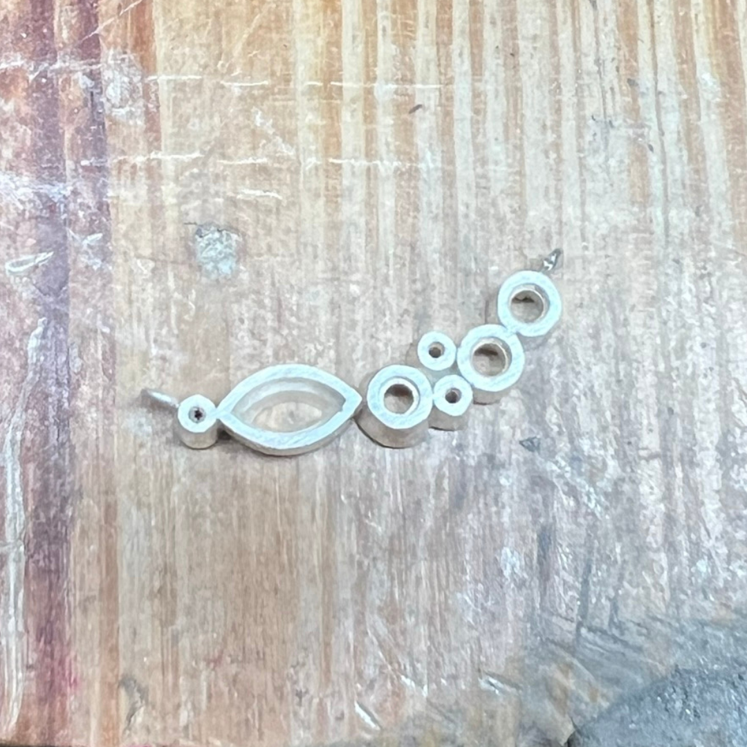 An unfinished necklace in the making, crafted in silver, showing the open bezels and framework for setting marquise-cut and round gemstones, laid on a wooden jewellery bench.